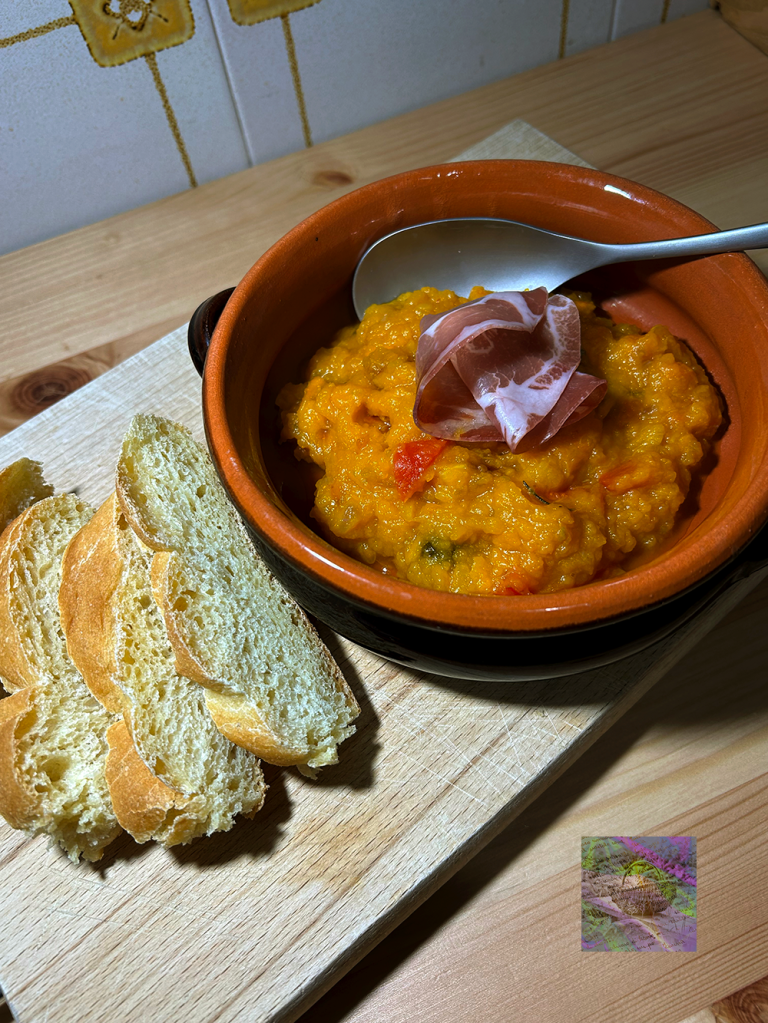 A bowl of creamy pumpkin soup garnished with a slice of ham, accompanied by slices of crusty bread, placed on a wooden cutting board.