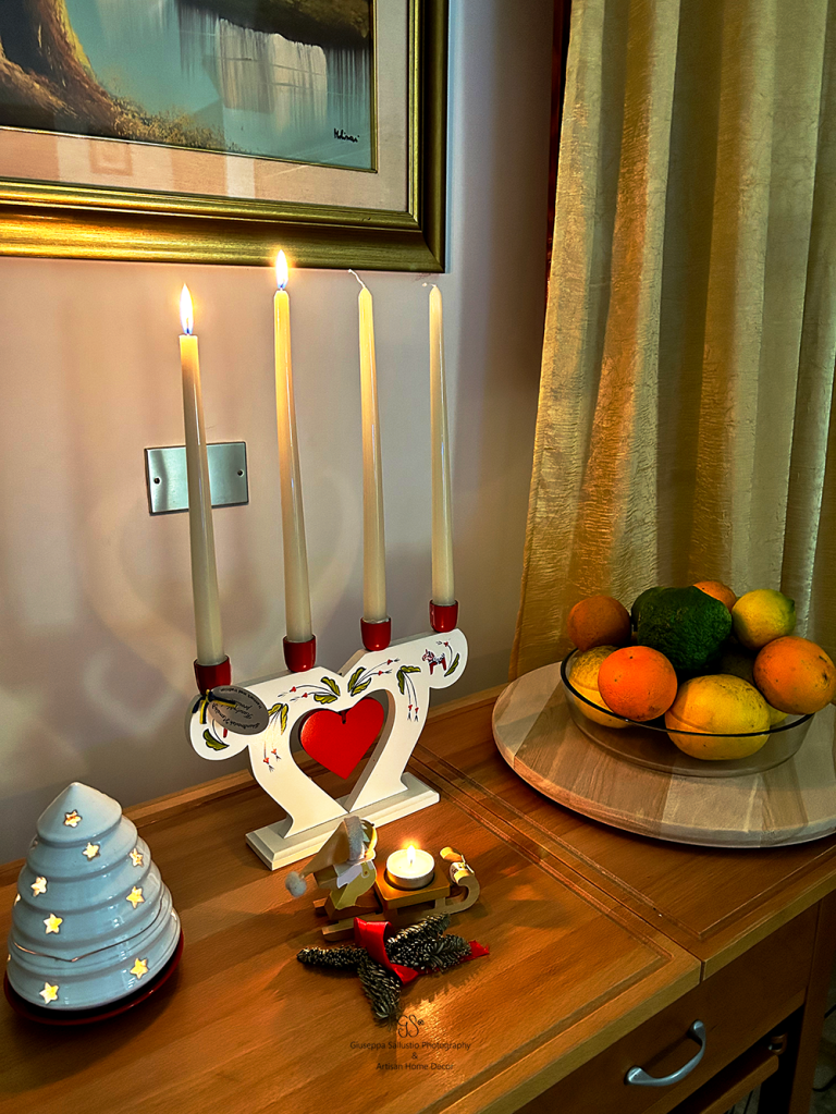 A cozy holiday scene featuring four lit candles arranged in a decorative holder with a heart design. In the foreground, a small candle illuminates a festive decoration of a wooden sled and pinecones. A white ceramic Christmas tree sits beside it, and a bowl of assorted fruits, including oranges and avocados, is displayed in the background.