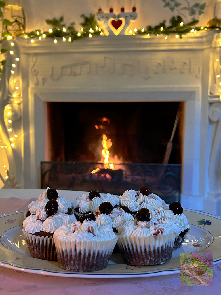 En tallrik med cupcakes dekorerade med grädde och körsbär, framför en brinnande eldstad med ljusslingor och musikinstrument på spiselkransen.