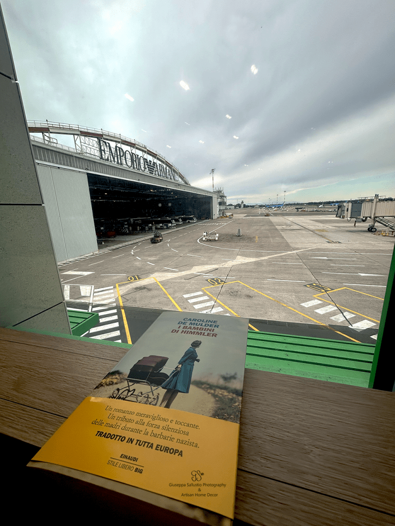 A view from inside Linate airport in Milan, showing a book titled 'I bambini di Himmler' by Caroline de Mulder resting on a table with the airport runway visible in the background.
