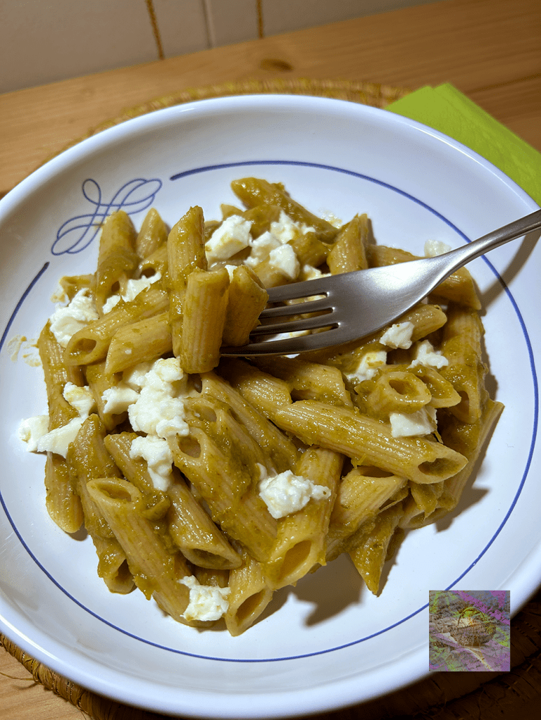 A bowl of whole wheat penne pasta mixed with pesto sauce and topped with crumbled feta cheese, accompanied by a fork.